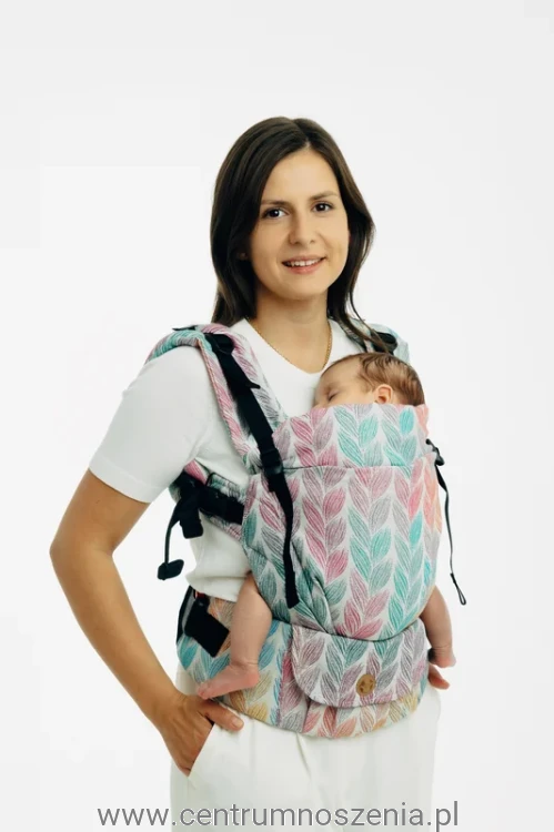 Mama nosząca niemowlę w ergonomicznym nosidle klamrowym w pastelowy wzór liści.